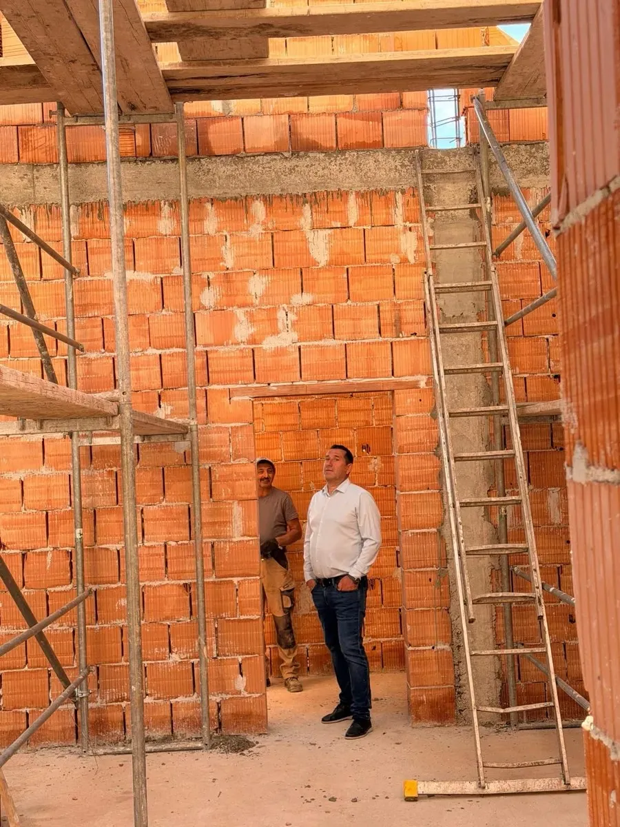 Doi bărbați stau într-o clădire din cărămidă netencuită, lângă schele metalice și o scară, sub grinzi din lemn.
