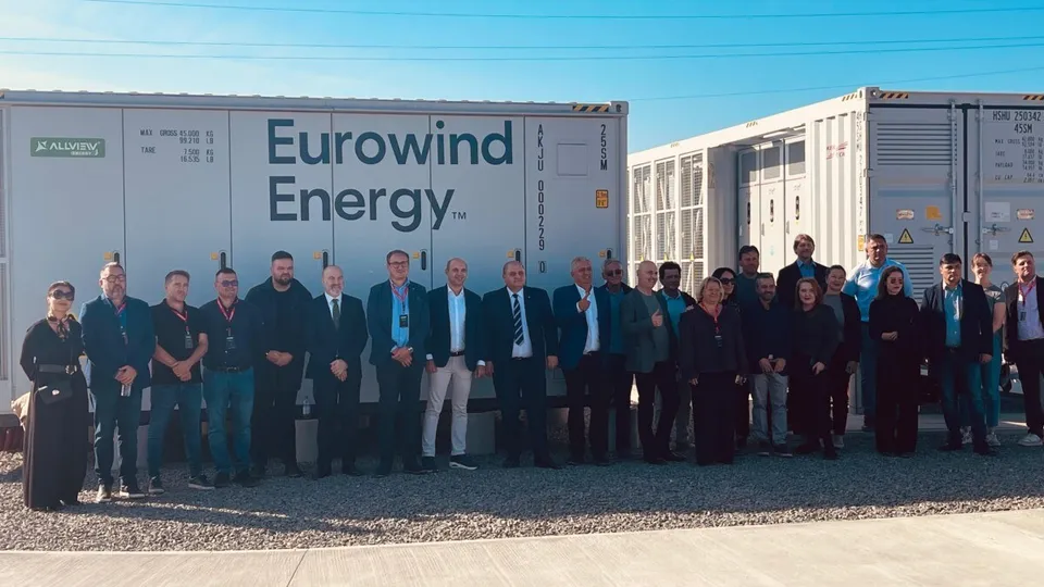 Un grup de persoane stă pe pietriș în fața unor containere industriale albe, unul inscripționat „Eurowind Energy”, sub cabluri aeriene.