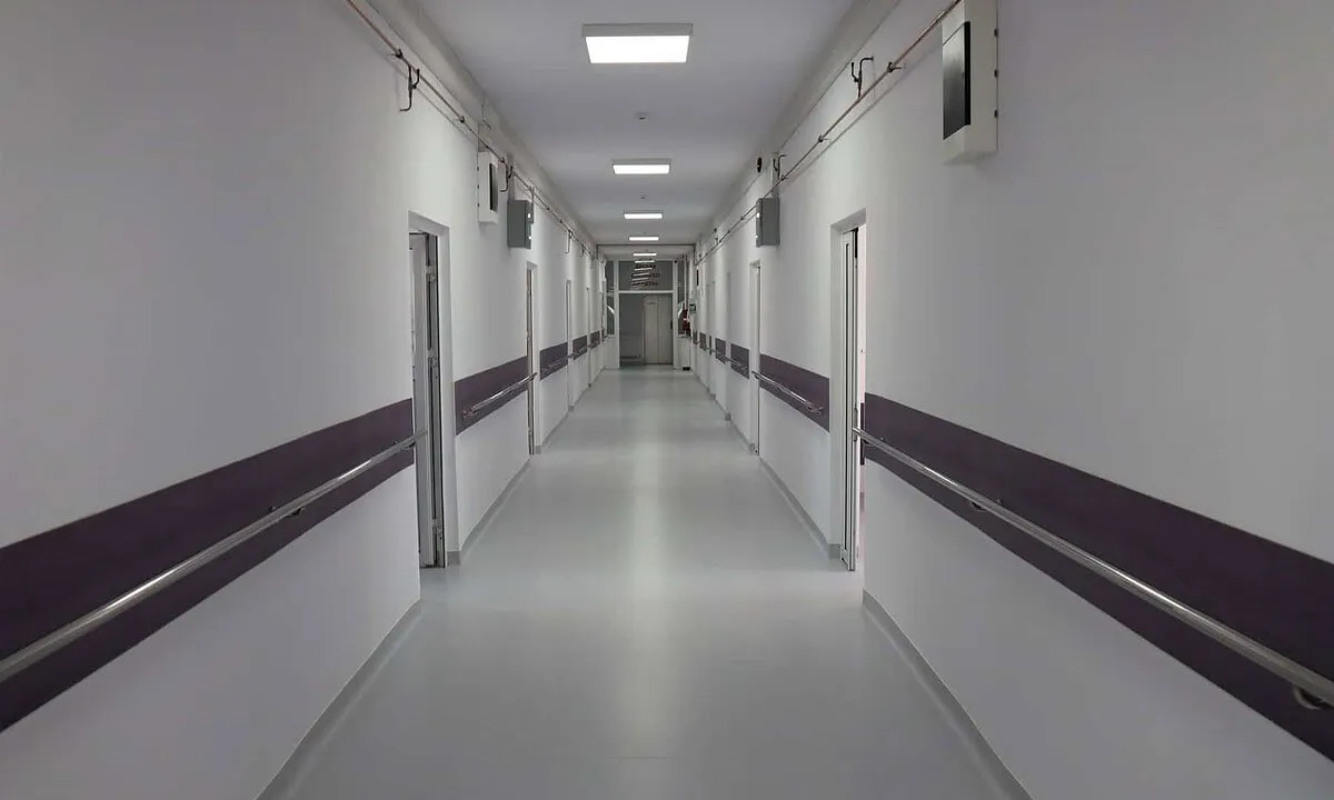 Un coridor lung, cu pereți albi, benzi închise la culoare, uși laterale și lumini dreptunghiulare în tavan.
