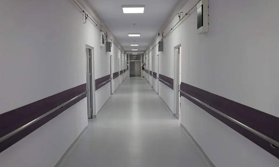 Un coridor lung, cu pereți albi, benzi închise la culoare, uși laterale și lumini dreptunghiulare în tavan.