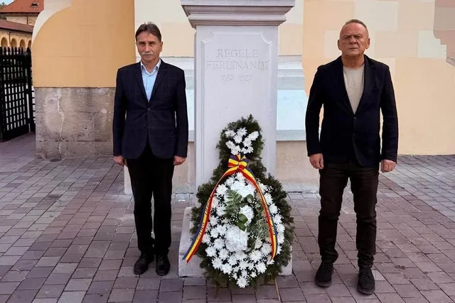 Doi bărbați în sacouri închise la culoare stau de o parte și de alta a unei coroane de flori albe, în fața unui monument.