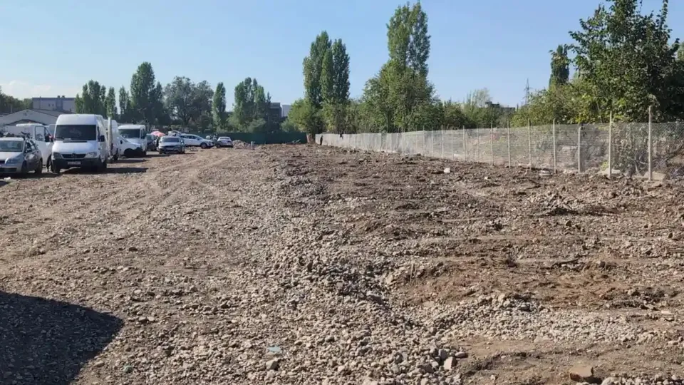 Un teren întins cu pietriș are mai multe vehicule parcate în stânga, un gard în dreapta și copaci în fundal.