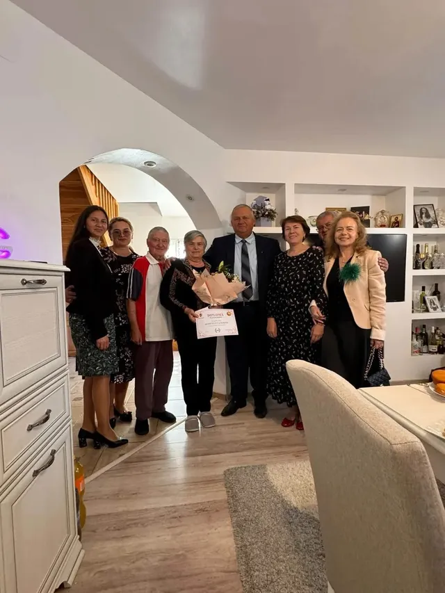 Șapte persoane stau într-o încăpere; două dintre ele țin un buchet de flori și o diplomă, iar mai multe poartă haine elegante.