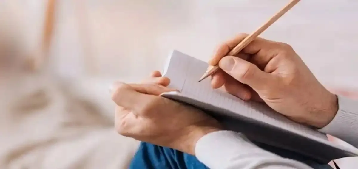 O persoană ține un carnet deschis și scrie cu un creion; în prim-plan se văd mâinile și mânecile deschise la culoare.