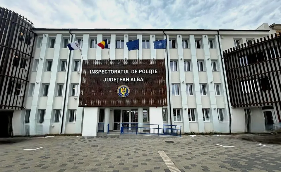 O clădire albă cu ferestre înalte are un panou inscripționat, mai multe steaguri la etaj și o curte pavată în față.