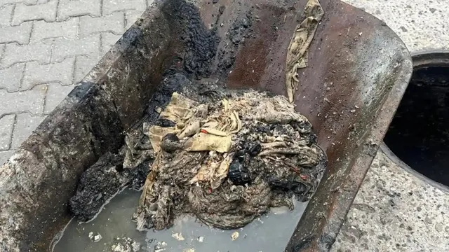 O lopată ține o grămadă de materiale îmbibate cu murdărie, lângă apă tulbure și o gură circulară întunecată.