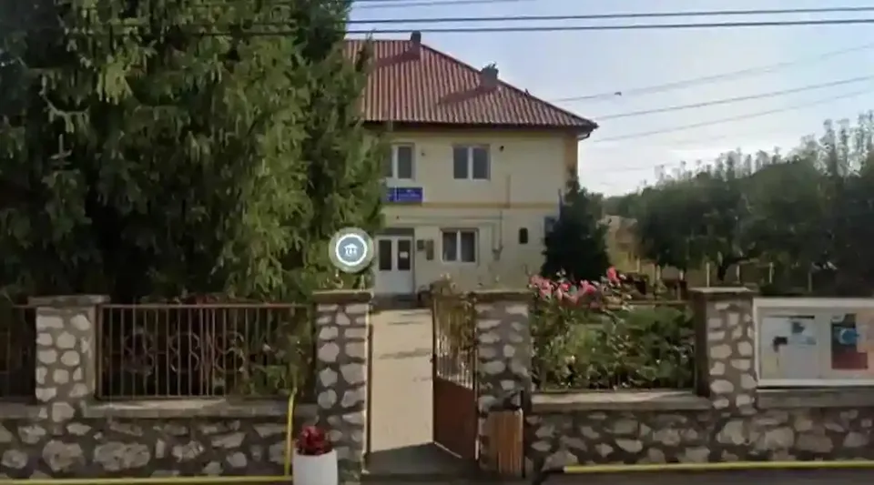 O clădire cu două niveluri și acoperiș roșu se află în spatele unui gard de piatră și metal, cu poarta deschisă.