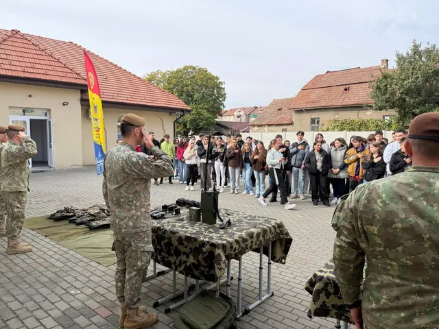 Doi militari în uniforme de camuflaj stau lângă mese acoperite cu pânză camuflată și echipamente, în fața unui grup de tineri.