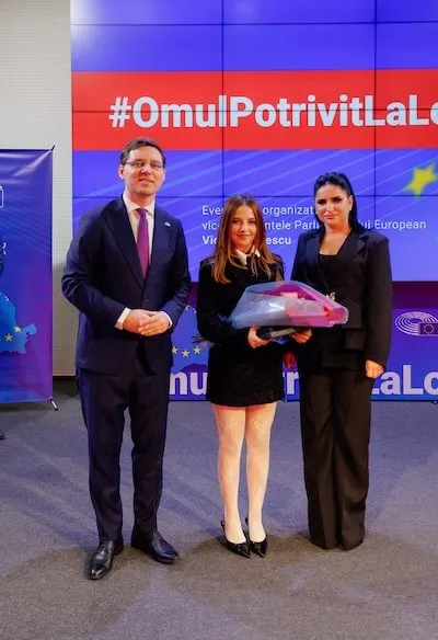 Trei persoane stau în fața unui panou colorat cu text; persoana din mijloc ține un buchet ambalat în folie roz.
