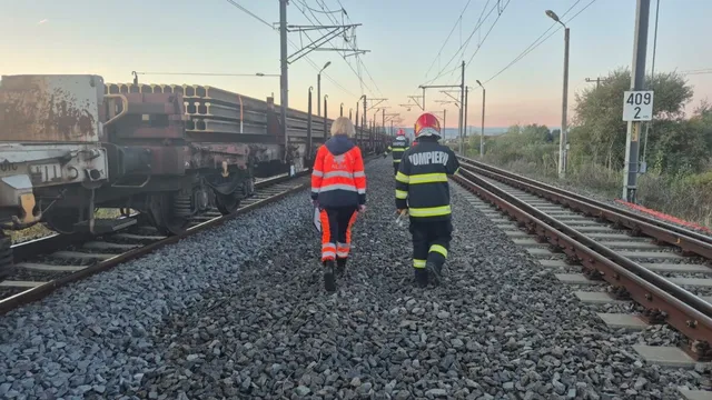 Două persoane în uniforme reflectorizante merg între șine, lângă vagoane de marfă, sub fire electrice și stâlpi de iluminat.