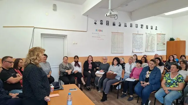 Într-o sală, mai multe persoane stau așezate pe scaune, iar o femeie stă lângă o masă pe care se află foi și o sticlă.
