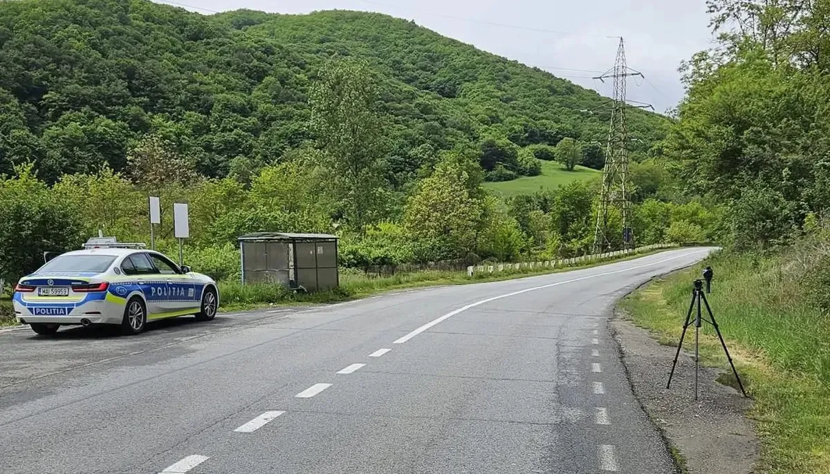 Un autoturism inscripționat staționează lângă un drum, iar un dispozitiv mic pe trepied se află pe marginea opusă, în fața dealurilor împădurite.