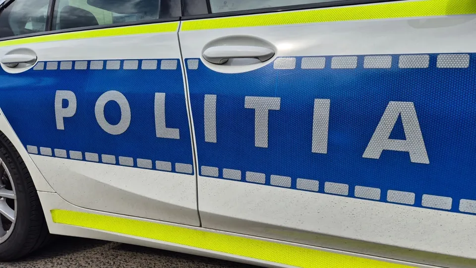 Prim-plan lateral al unei mașini albe, cu bandă albastră inscripționată „POLIȚIA” și elemente reflectorizante galbene.