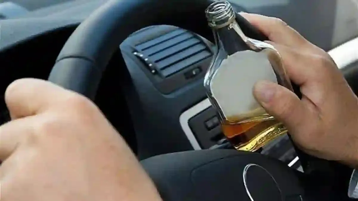 Două mâini se află lângă un volan, iar una ține o sticlă deschisă cu lichid gălbui.
