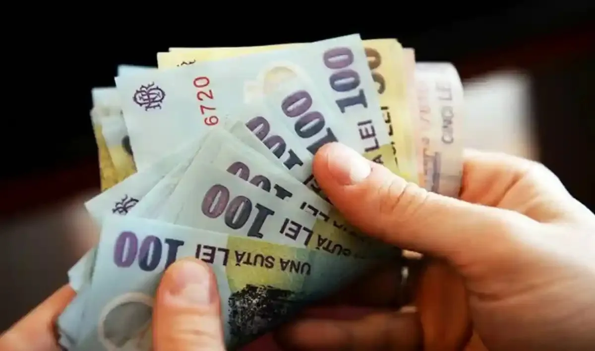 Două mâini țin în evantai mai multe bancnote de 100 de lei, pe un fundal întunecat.