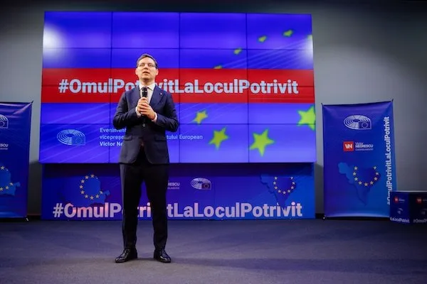 Un bărbat în costum închis la culoare ține un microfon pe o scenă, în fața unui ecran cu text și stele colorate.