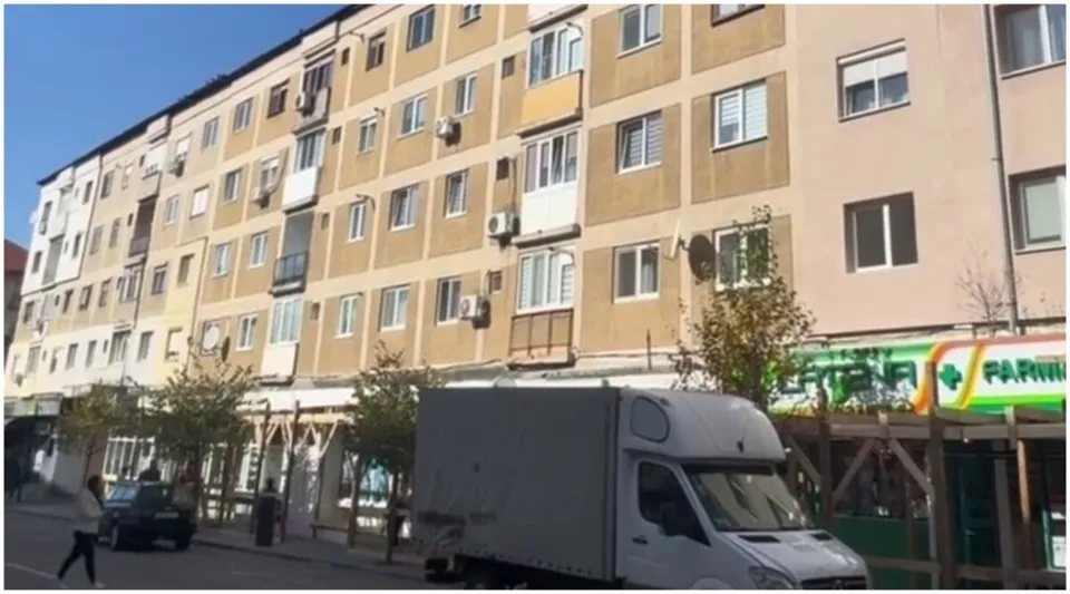 Un bloc cu mai multe etaje are fațadă bej; la parter se văd structuri din lemn, iar în față se află o autoutilitară albă.
