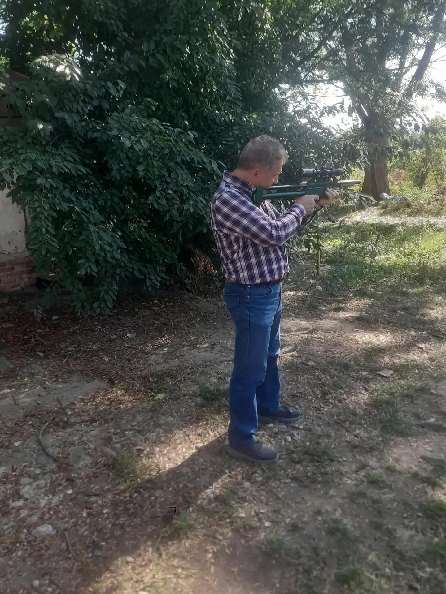 Un bărbat în cămașă în carouri și blugi ține la umăr o armă lungă, într-o zonă cu arbori și pământ.