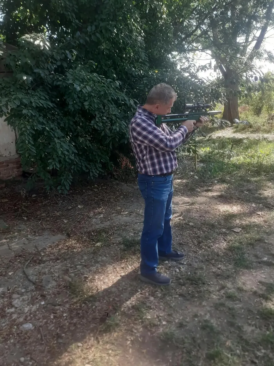 Un bărbat în cămașă în carouri și blugi ține la umăr o armă lungă, într-o zonă cu arbori și pământ.