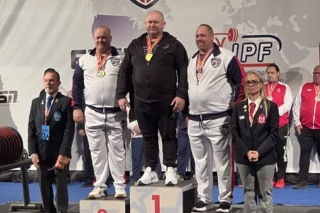 Trei bărbați cu medalii la gât stau pe podium, purtând treninguri albe și negre; alte persoane se află lângă ei.