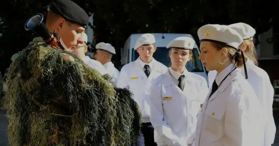O persoană în uniformă verde, acoperită cu material vegetal, stă în fața mai multor tineri îmbrăcați în uniforme albe și purtând berete.