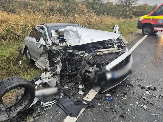 Un autoturism argintiu are partea din față grav avariată; pe carosabil sunt împrăștiate fragmente, iar în fundal se vede parțial un vehicul roșu.