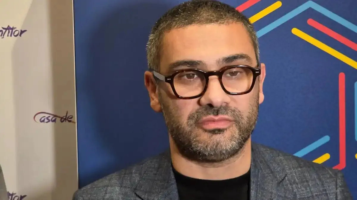 Un bărbat cu ochelari negri, barbă scurtă și sacou gri se află în fața unui panou albastru cu linii colorate.