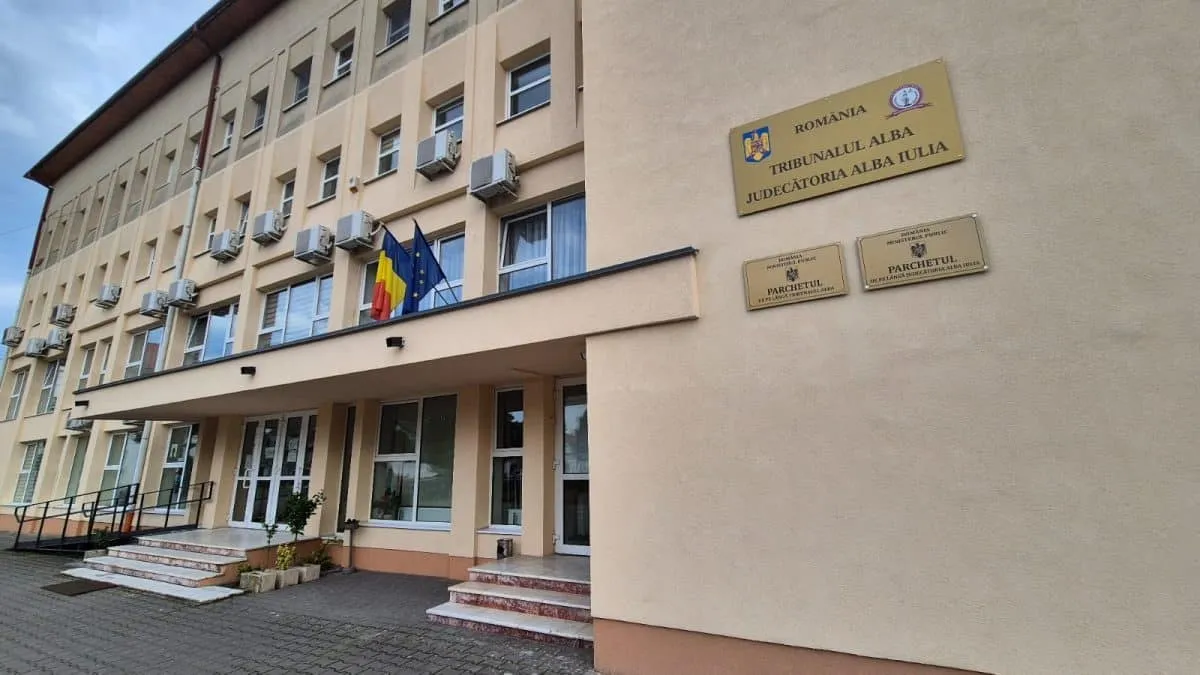 O clădire bej cu ferestre, intrare cu trepte, trei steaguri și plăcuțe inscripționate pe fațadă.