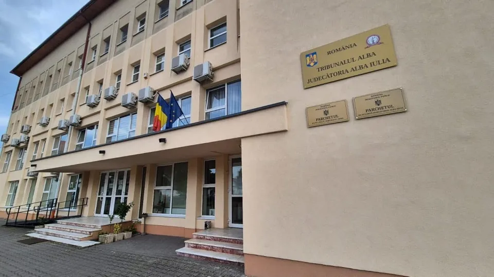 O clădire bej cu ferestre, intrare cu trepte, trei steaguri și plăcuțe inscripționate pe fațadă.