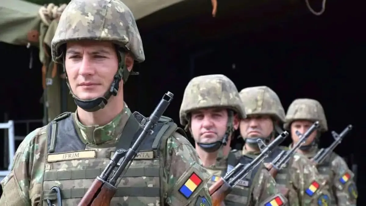 Mai mulți militari în uniforme de camuflaj și căști stau unul în spatele altuia; în față se văd arme și ecusoane colorate.