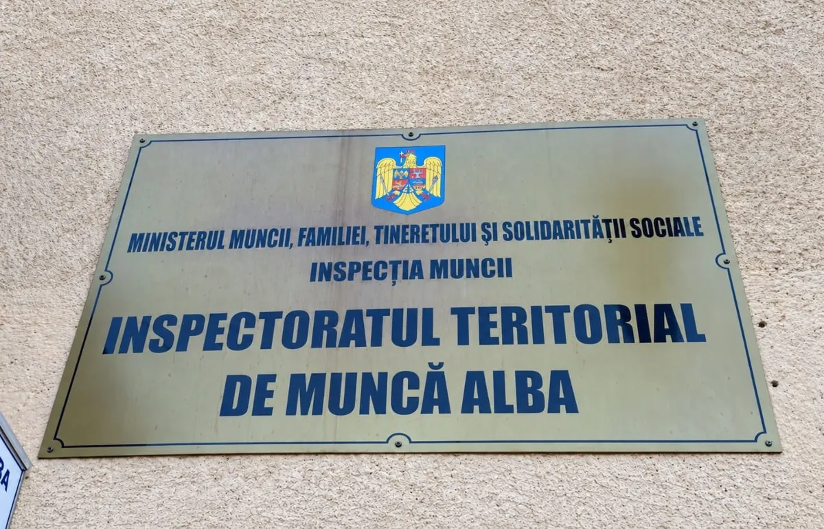 O plăcuță dreptunghiulară cu text albastru și o stemă colorată este fixată pe un perete deschis la culoare.