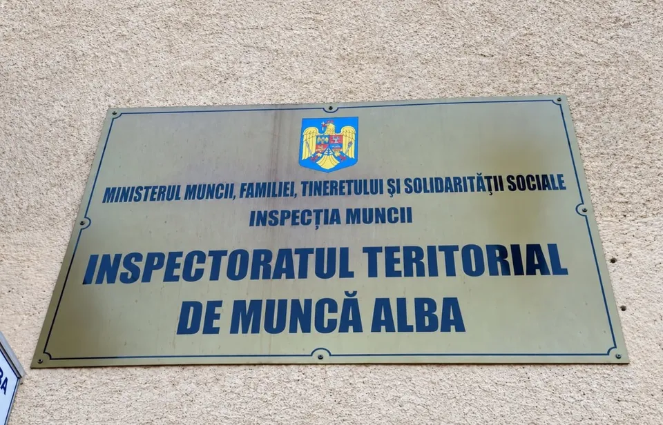 O plăcuță dreptunghiulară cu text albastru și o stemă colorată este fixată pe un perete deschis la culoare.