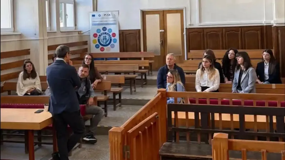 Un bărbat în sacou vorbește în fața mai multor persoane așezate pe bănci de lemn, într-o sală cu lambriuri.