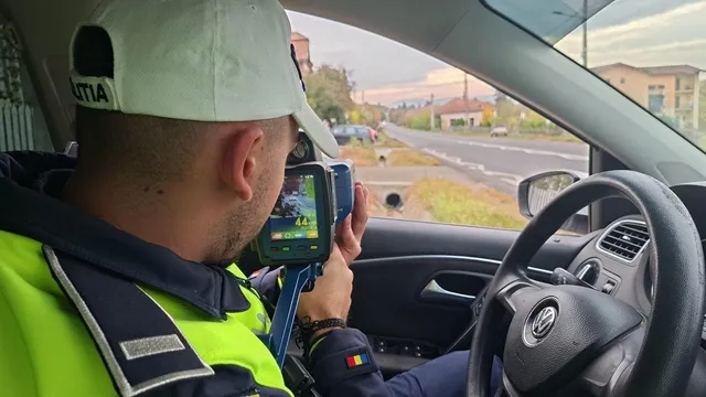 Un bărbat cu șapcă și vestă reflectorizantă stă într-un autoturism și ține un dispozitiv cu ecran; prin geam se vede o șosea.