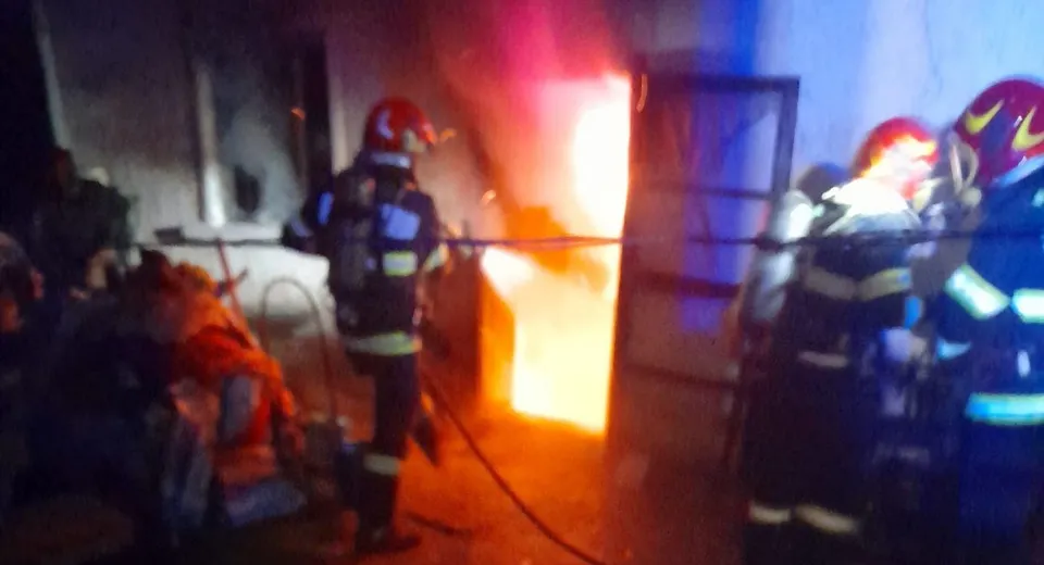 Mai multe persoane în echipament reflectorizant, inclusiv una ghemuită în stânga, se află lângă o intrare cu flăcări și fum.