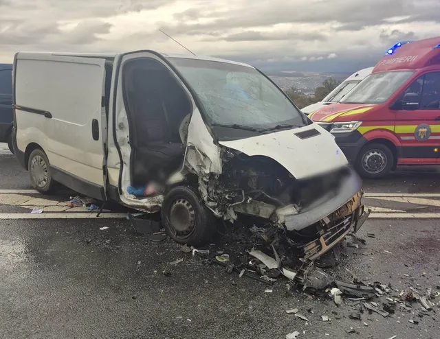 O dubă albă are partea frontală grav avariată, portiera laterală deschisă și resturi împrăștiate pe șosea; în spate se află o ambulanță roșie.