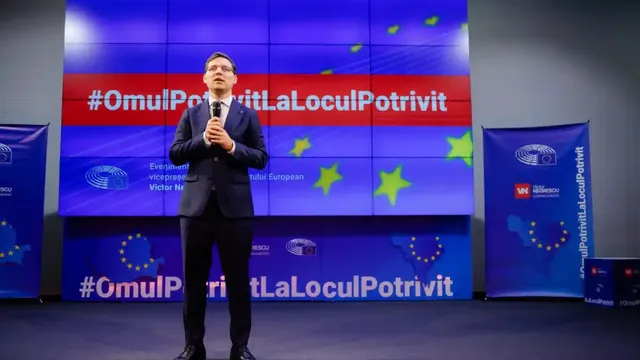 Un bărbat în costum închis la culoare stă pe o scenă și ține un microfon; în spatele lui sunt ecrane albastre cu text.