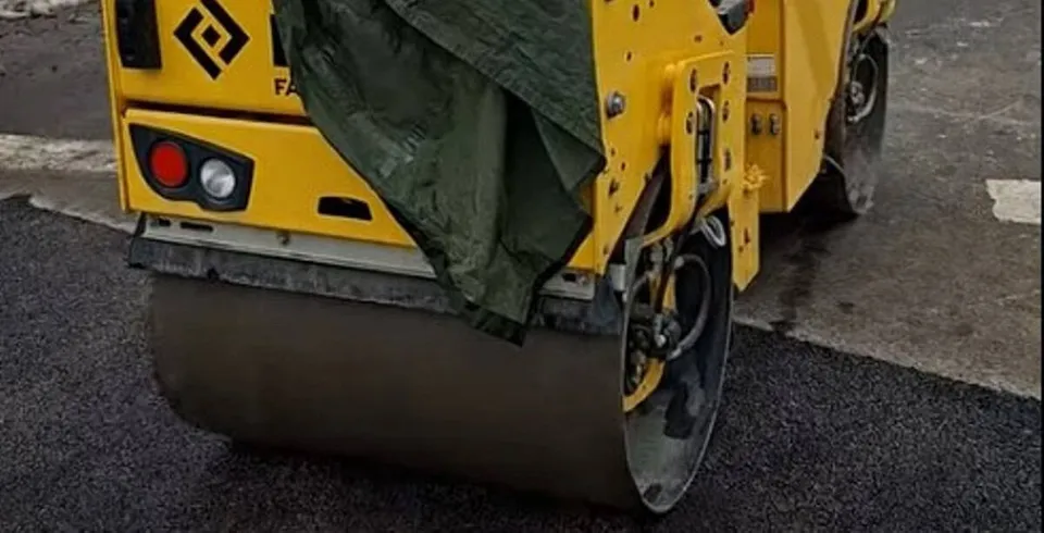 Un utilaj galben cu cilindru metalic se află pe un strat de asfalt închis la culoare; o prelată întunecată îi acoperă parțial partea superioară.