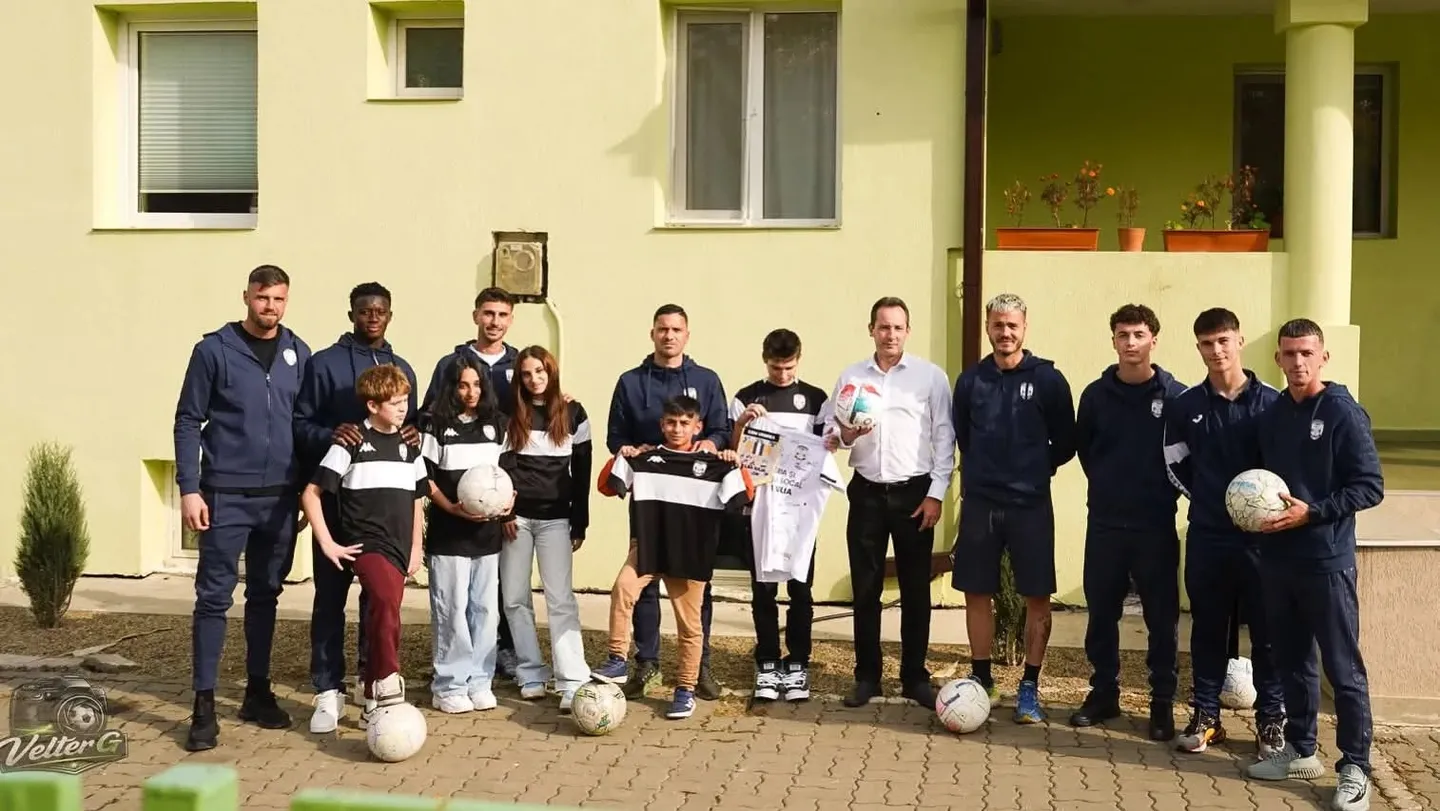 Mai mulți adulți și copii stau în fața unei clădiri; unii poartă echipament sportiv și țin mingi și tricouri albe.