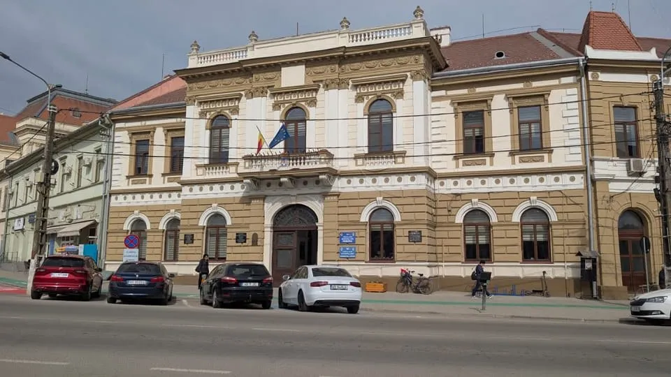 O clădire cu fațadă deschisă la culoare, ferestre arcuite și steaguri la balcon se află în spatele mai multor mașini și pietoni.