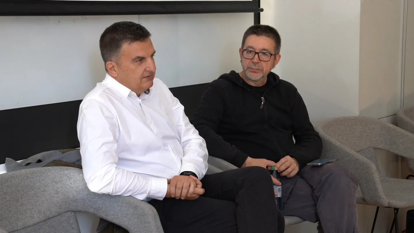 Doi bărbați stau pe fotolii gri; unul poartă o cămașă albă, iar celălalt poartă o bluză neagră și ochelari.