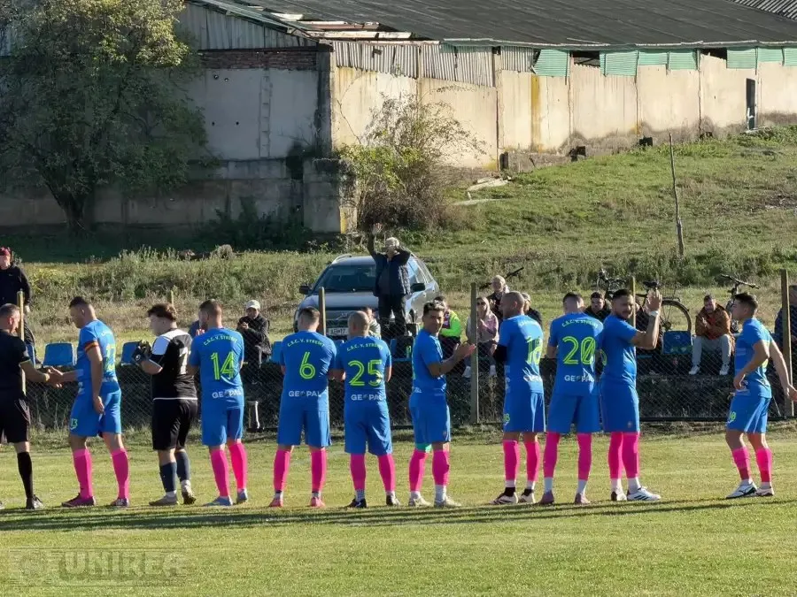 Fotbaliști în echipamente albastre și negre stau în șir pe terenul cu iarbă; în spatele lor se văd câteva persoane și o mașină.