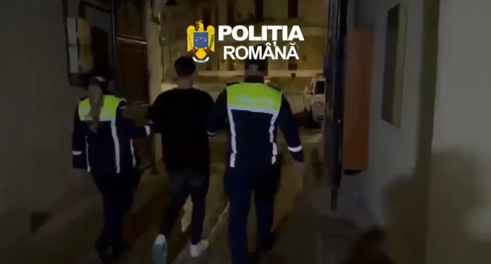 Doi bărbați în veste reflectorizante însoțesc un bărbat îmbrăcat în negru pe un coridor slab luminat.