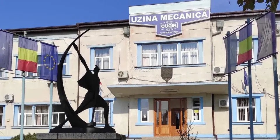 O clădire deschisă la culoare are ferestre, o intrare cu uși din lemn și o firmă deasupra; în față se află o sculptură și steaguri.