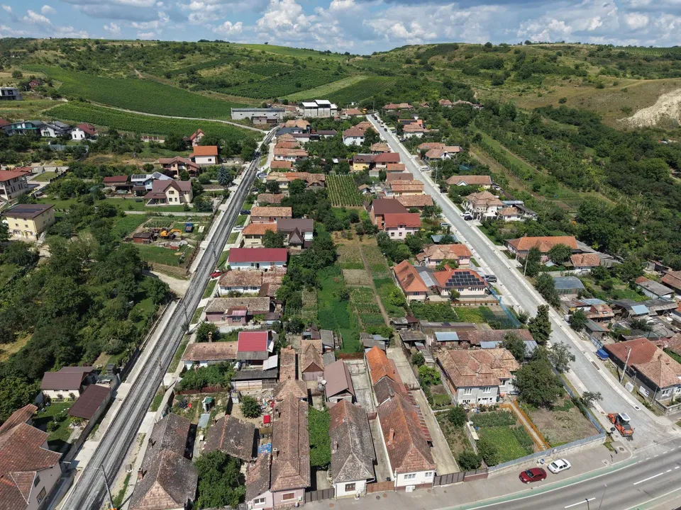 Vedere aeriană cu case și acoperișuri roșiatice, grădini și două drumuri lungi între clădiri, pe un fundal de dealuri verzi.
