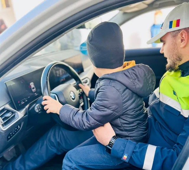 Un copil cu căciulă neagră stă la volanul unei mașini, lângă un bărbat cu șapcă albă și jachetă albastră.