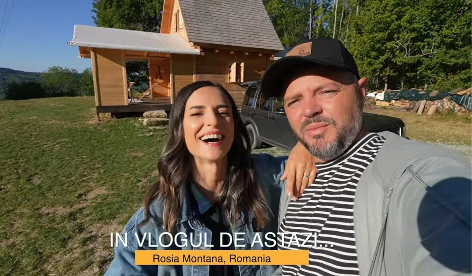 O femeie cu jachetă albastră și un bărbat cu șapcă neagră stau în prim-plan, lângă o mașină, cu o casă și copaci în fundal.
