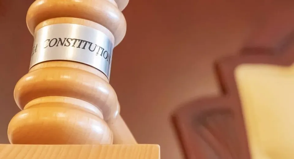 Un ciocan de lemn ocupă prim-planul, cu o bandă albă inscripționată; în fundal se văd suprafețe maro și o zonă galbenă.