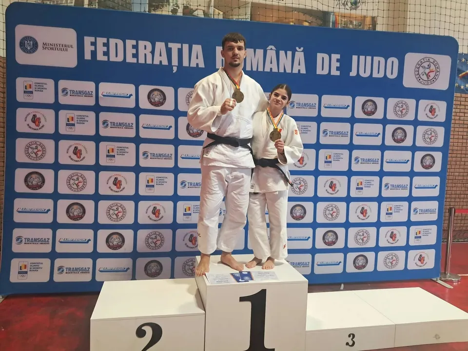 Două persoane în echipamente albe de judo, cu centuri negre și medalii la gât, stau pe un podium în fața unui panou cu sigle.