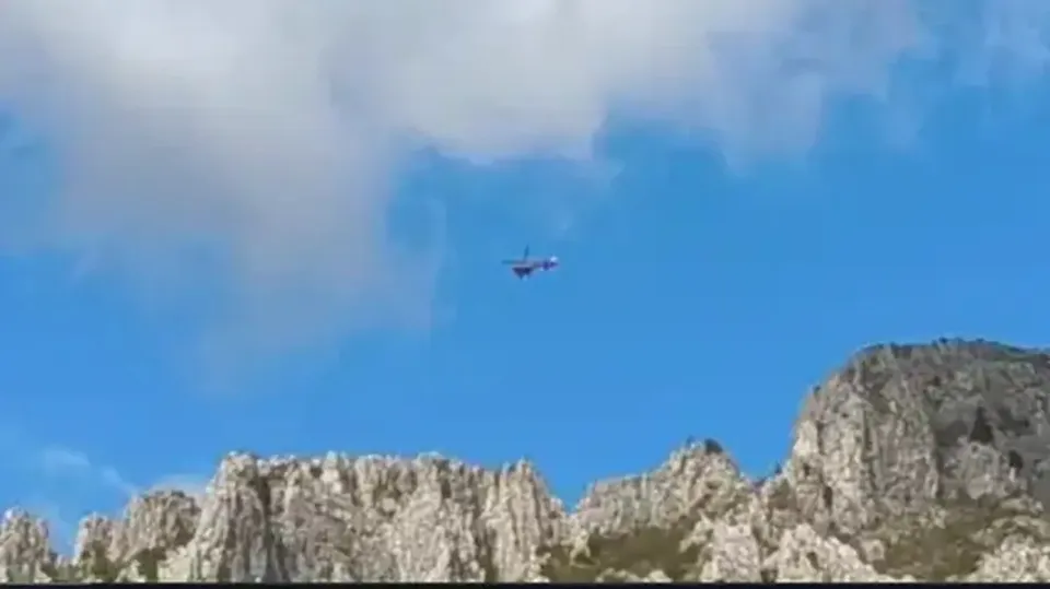 Un elicopter mic zboară pe un cer albastru deasupra unor stânci deschise la culoare.